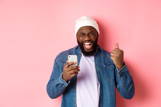 Technology and online shopping concept. Happy Black man rejoicing, winning in app, holding smartphone and scream yes, standing over pink background.