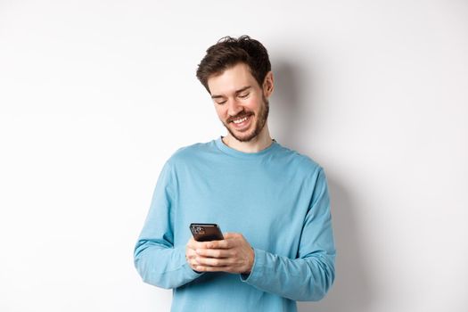 Portrait of modern caucasian man with beard, wearing casual clothes, reading smartphone screen and smiling, networking via mobile phone, standing over white background.