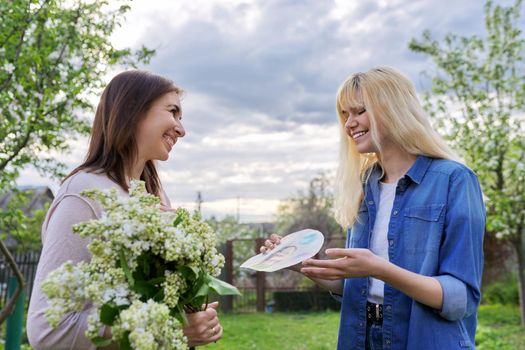 Mother's Day, female teenager congratulating her mother with bouquet of flowers and picture. Mom and daughter embracing, in backyard, outdoor. Family, holiday, relationship, love, happiness