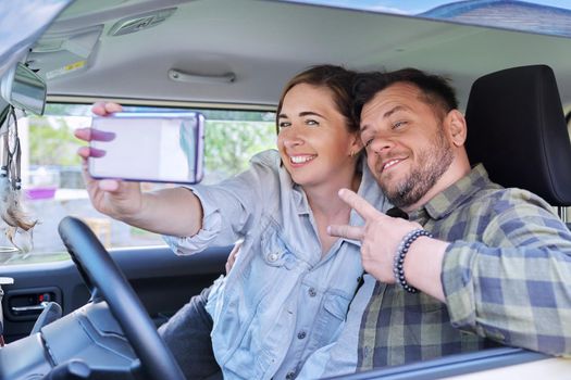 Happy married couple taking selfie photo on smartphone, husband and wife sitting in car.