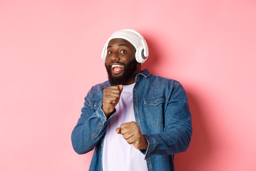 Happy african-american man dancing, listening music in headphones and looking at camera, standing over pink background.