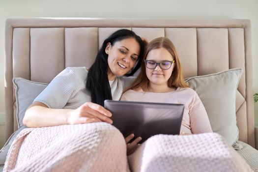 Mother and teenage daughter resting at home together, use digital tablet. Woman and teen girl lying in bed, looking at screen, movie video. Family, parent teeneger relationship, vacation, technology