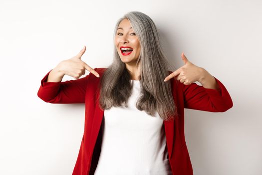 Ambitious senior woman pointing fingers at center, showing logo and smiling, standing over white background.