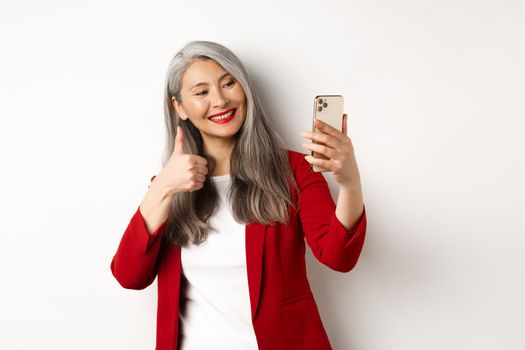 Successful asian businesswoman in red blazer, taking selfie on smartphone with thumb-up, showing approval, standing over white background.