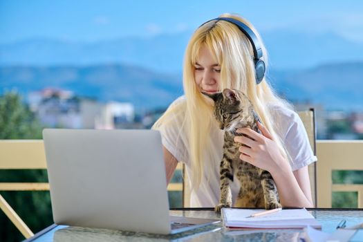 Female student studies at home online using laptop. Teenager sitting on outdoor balcony with pet cat in headphones, school notebooks, looking at screen. E-learning, modern technologies in education