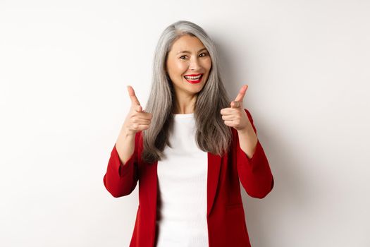 Professional female employer in trendy red blazer and makeup, pointing fingers at camera and smiling, praising something, need you, standing over white background.