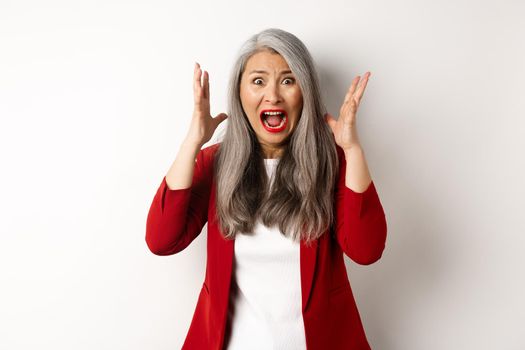 Asian senior businesswoman screaming and looking outraged, feeling distressed and shouting, standing against white background.