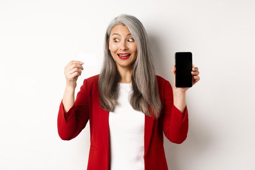 Cheerful asian mature woman showing blank smartphone screen and credit card, concept of e-commerce.