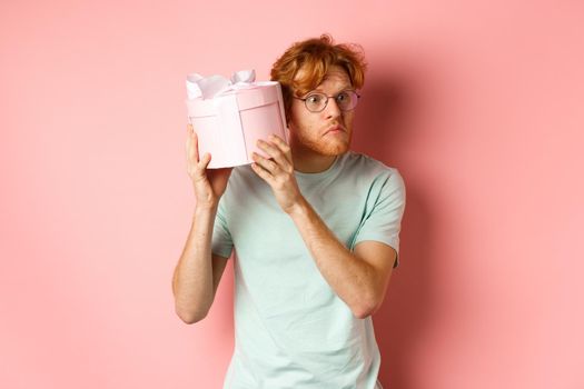 Love and holidays concept. Intrigued redhead guy press ear to box and shaking gift, guessing what inside, standing over pink background.