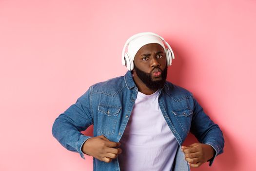 Cool and sassy Black guy dancing, listening to music in headphones, looking confident, standing over pink background. Copy space