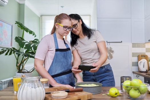 Mom and teenage daughter preparing apple pie together, at home in the kitchen. Woman photographing pie on smartphone. Family, parent teenager relationship, lifestyle, eating at home concept