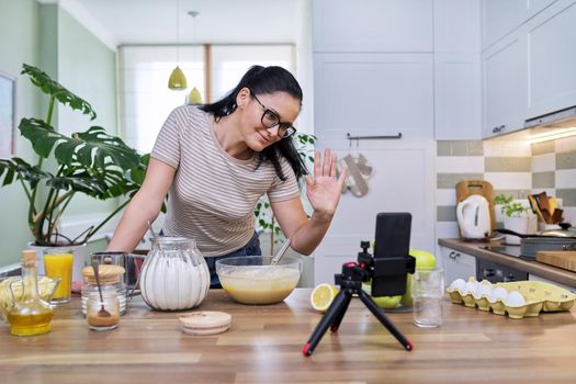 Technologies in everyday life, 40s woman preparing apple pie at home in kitchen, with smartphone on tripod using video call for communication. Lifestyle, eating at home, technology, people concept