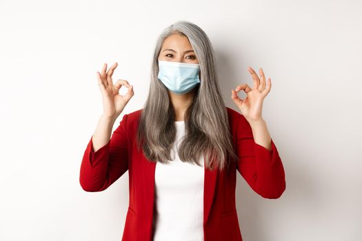 Coronavirus and business concept. Asian female manager in face mask looking cheerful, showing okay signs in approval, white background.