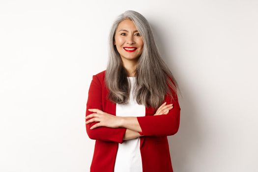 Business people. Smiling asian senior woman in red blazer, cross arms on chest and looking professional, standing over white background.