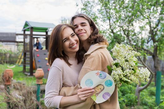Teenage son congratulating mom on Mother's Day with a bouquet of lilac flowers and a handicraft picture, outdoor in the backyard. Family, holiday, relationship, love, happiness