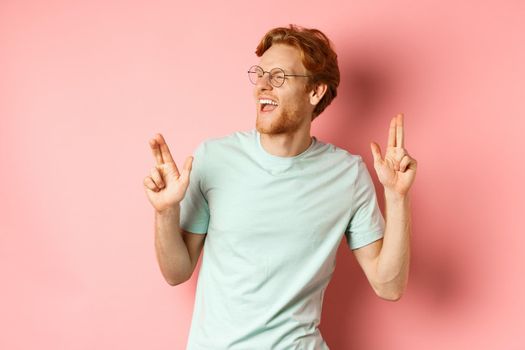 Cheerful redhead guy listening music and dancing, triumphing or celebrating success, standing over pink background.