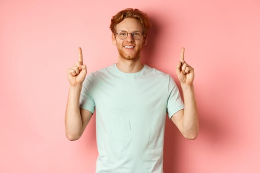 Attractive young man in glasses and t-shirt showing promo deal, pointing fingers up and smiling at camera, standing over pink background.