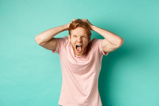 Angry redhead man screaming and ripping hair out of head, yelling and losing temper, looking insane from rage, standing over mint background.