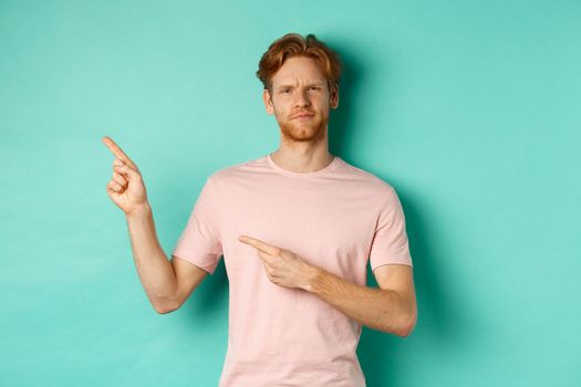 Skeptical and unamused redhead man frowning, pointing fingers at upper left corner banner, standing over turquoise background.