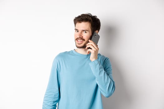 Technology concept. Young male model talking on mobile phone, calling someone on smartphone and smiling, standing over white background.