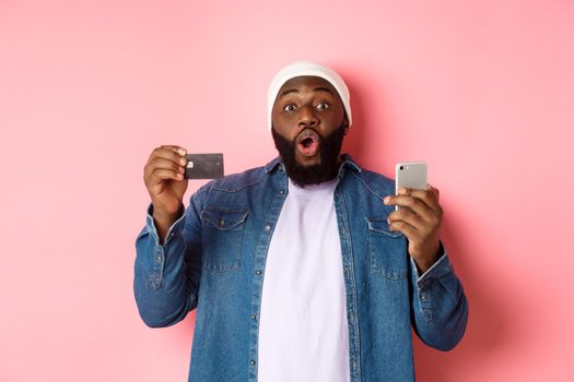 Online shopping. Excited Black man showing credit card, using smartphone, staring at camera amazed, standing over pink background.