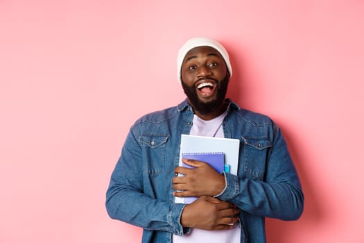 Education. Happy african-american male student in beanie holding notebooks, studying courses, smiling at camera, standing over pink background.