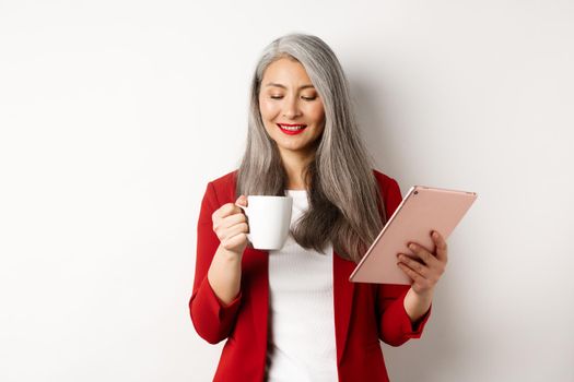 Business people concept. Asian mature businesswoman holding digital tablet and looking at coffee cup with pleased smile, standing over white background.