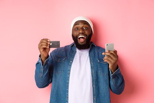 Online shopping. Happy bearded african-american man smiling, showing credit card and making purchase on smartphone, standing over pink background.