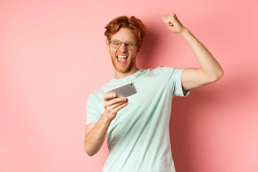 Happy redhead man winning in mobile video game, raising hand up and shouting yes with joy, celebrating victory, looking at smartphone, standing over pink background.