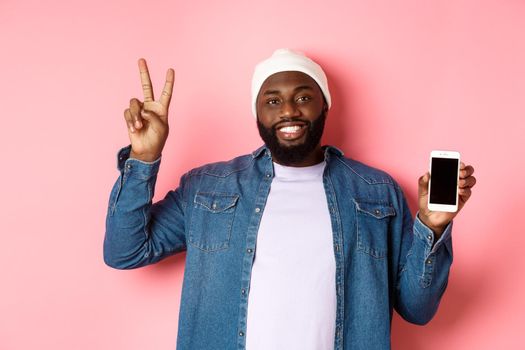 Online shopping and technology concept. Happy Black guy in beanie and denim shirt showing mobile screen and peace sign, standing over pink background.