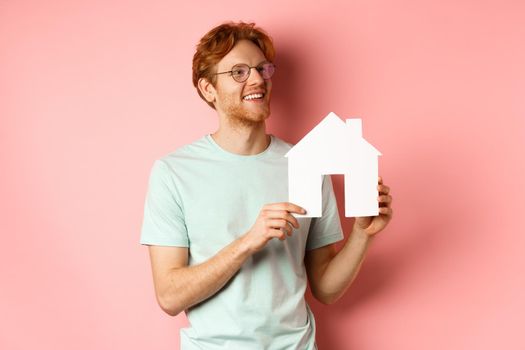 Real estate. Happy redhead man dreaming of buying property, looking right and showing paper house cutout, standing over pink background.