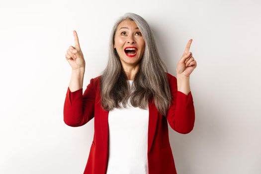 Happy asian lady in red blazer and makeup, looking and pointing fingers up, showing special offer, standing over white background.