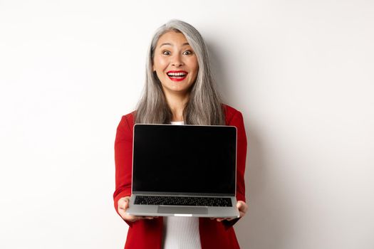 Business. Happy senior female entrepreneur demonstrate blank laptop screen, smiling amazed at camera, standing over white background.