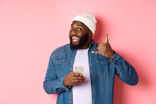 Happy Black man showing call me sign, making phone gesture and smiling, holding smartphone, standing in beanie and denim shirt over pink background.