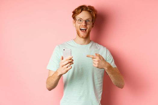 Cheerful guy talking about internet promo, smiling amazed and pointing finger at smartphone, standing over pink background.