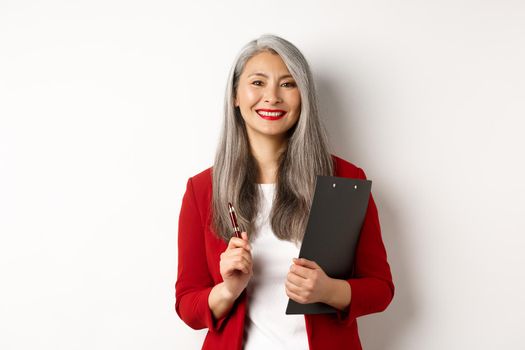 Successful asian lady boss in red blazer, holding clipboard with documens and pen, working and looking happy, white background.