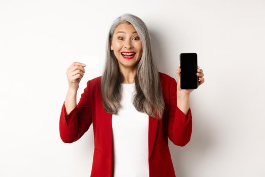 Cheerful asian mature woman showing blank smartphone screen and credit card, concept of e-commerce.