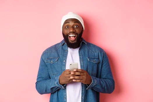 Technology and online shopping concept. Surprised young Black man using mobile phone, looking at camera amazed and happy after reading message, pink background.