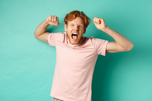 Redhead male fan watching sport game and cheering, shouting and making fist pumps, looking at camera, watching competition, rooting for team, standing over turquoise background.