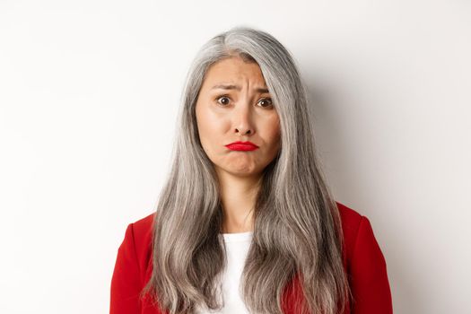 Close up of sad and unhappy asian senior woman sulking, frowning and looking jealous at camera, standing over white background.