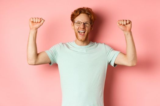 Happy winner with red hair and beard celebrating victory, shouting yes with joy and raising hands up, enjoy success, standing over pink background.
