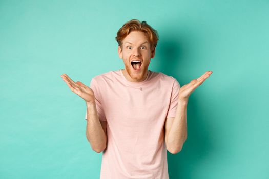 Portrait of surprised and happy young man with red hair, spread hands sideways and gasping with awe at camera, looking amazed, standing over turquoise background.