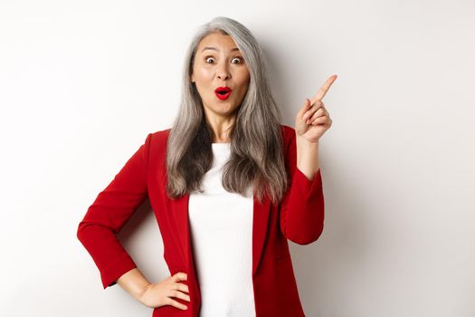 Amazed senior asian lady pointing upper right corner, staring excited at camera, checking out promo offer, standing over white background.