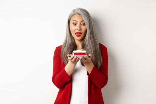 Image of funny asian senior woman looking tempted at piece of cake, desire to bite dessert, standing over white background.