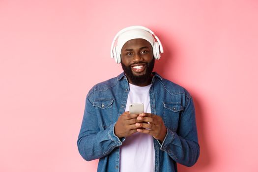 Image of handsome Black guy in headphones, listening music and using mobile phone, smiling at camera, pink background.