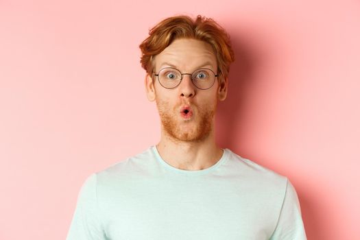 Close up of impressed redhead man in glasses saying wow, raising eyebrows surprised and staring at camera, standing over pink background.