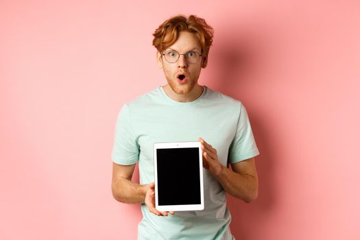 Excited young man with red hair and beard, checking out online promotion, showing digital tablet screen and staring at camera amazed, standing over pink background.