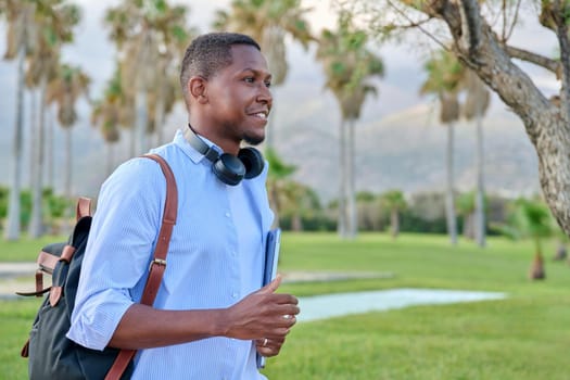 Outdoor portrait of young man with laptop headphones and backpack. African male adult student, creative freelancer, businessmen in tropical park. E-learning, online technologies, education, business