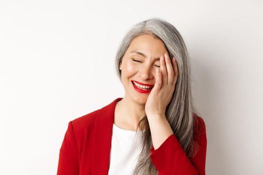 Beauty and makeup concept. Close up of beautiful asian mature woman, laughing and touching face, smiling happy, standing over white background.