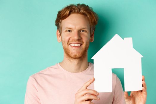 Real estate concept. Close up of redhead bearded man in t-shirt showing paper house cutout, smiling happy at camera, standing over turquoise background.